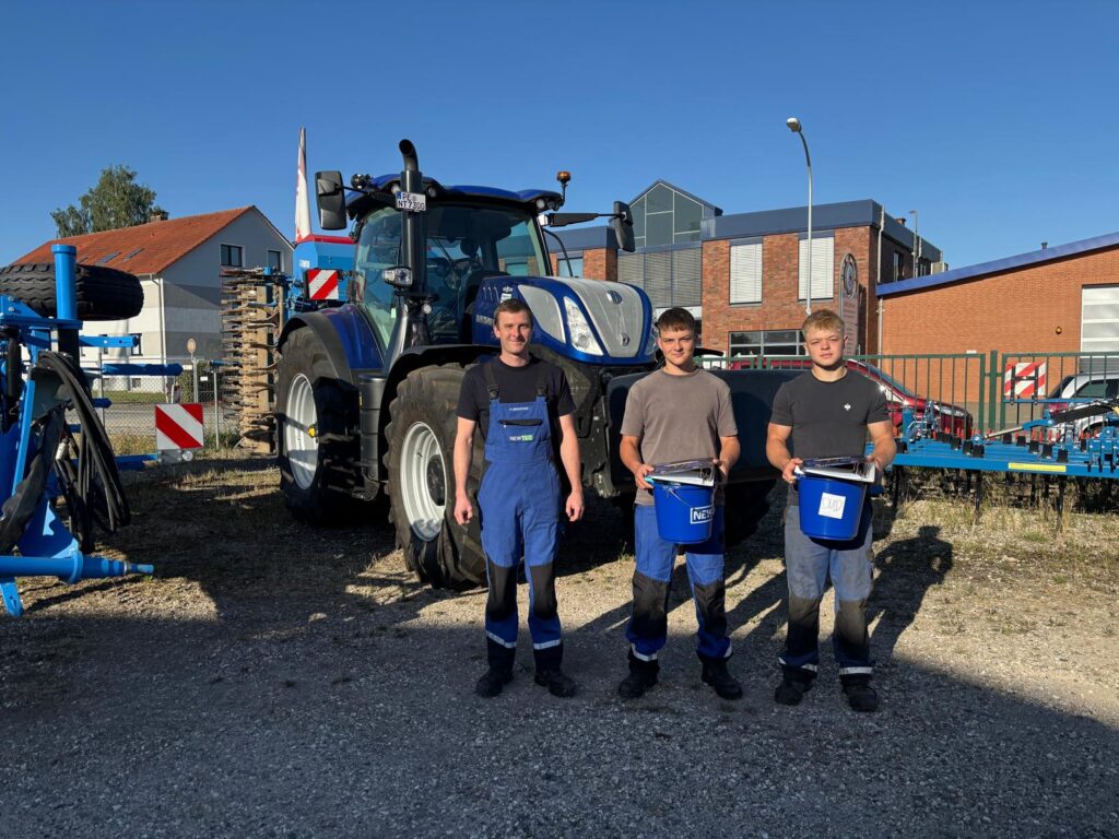Drei junge Männer in Arbeitskleidung stehen vor einem blauen Traktor und halten blaue Eimer in der Hand