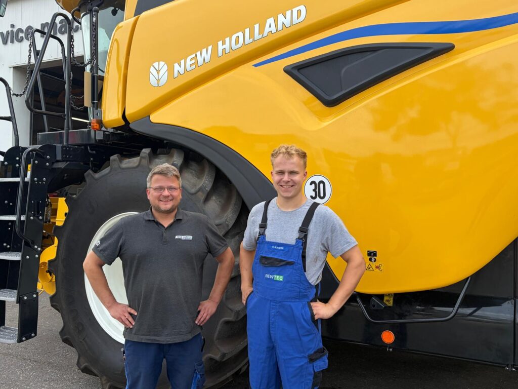Zwei Männer stehen vor einem gelben New-Holland-Mähdrescher, einer trägt blaue Arbeitslatzhose, der andere ein schwarzes Poloshirt