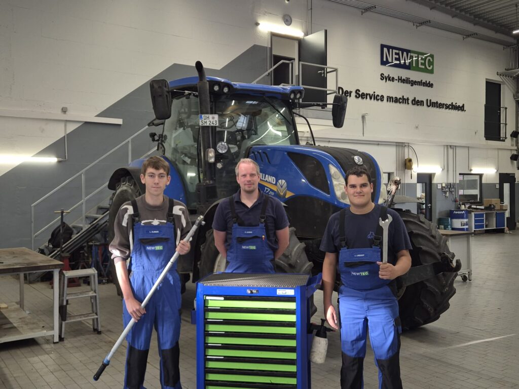 Drei Männer in blauen Arbeitslatzhosen stehen vor einem großen blauen Traktor in einer Werkstatt, zwei halten Werkzeuge, im Hintergrund ein Schild mit der Aufschrift 'NEWTEC Syke-Heiligenfelde'